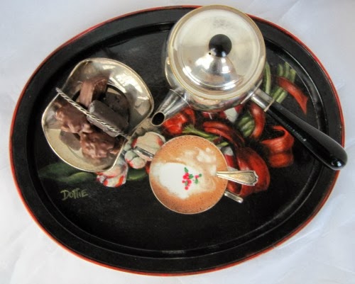 overhead view of a round dark serving tray with a colorful central design holding a cup of Wittamer  hot chocolate  topped with red and green sprinkles, a serving dish with chocolate candies and a silver hot chocolate pot.