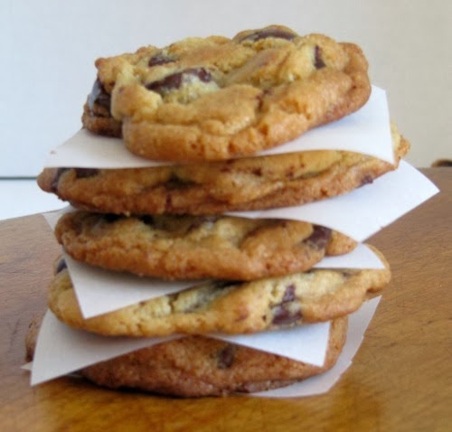 side view of a stack of  jacques torres 
 the best chocolate chip cookies separated by pieces of white parchment sitting on a brown surface.