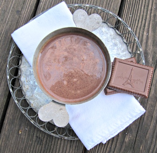 overhead view of a cup of chocolat chaud or French hot chocolate from dorie greenspan is sitting on a wire round serving tray on a wood surface along with a white napkin and two pieces of chcoclate candy