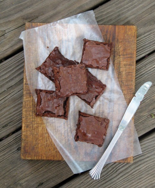Overhead view of a collection of chocolate fudge brownie squares are sitting on a piece of white parchment along with a sliver butter knife on a wooden surface