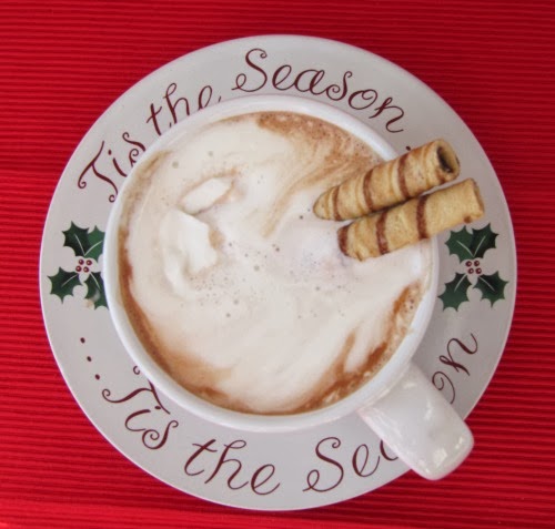 overhead view of a white suacer decorated with a red christmas message and holly berries. The cup holds a generous serving of Hershey's "Perfectly Chocolate" Hot Cocoa topped with whipped creamand garnished with two rolled wafer cookies. The cup and saucer is on a red background.