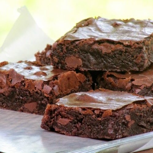 Front view of a pile of chocolate fudge brownie squares with crispy crinkled tops on a white paper on a wood cutting board