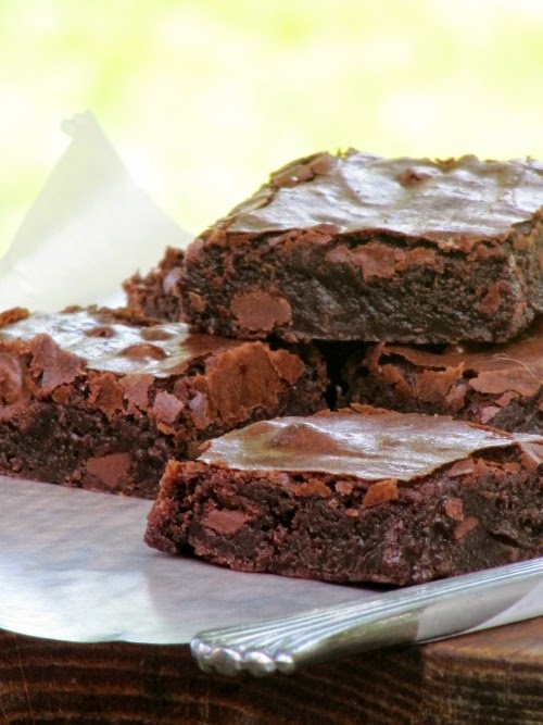 Front view of a pile of baked chocolate fudge brownies squares with crispy crinkled tops on a white paper on a wood cutting board.