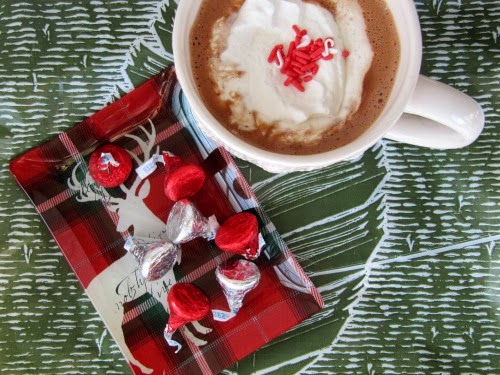 Overhead view of a white ceramic mug of creamy homemade hot chocolate topped with whipped cream and red candy sprinkles on a gray and white patterned cloth next to a red and white tray filled with silver and red wrapped hershey's kisses.
