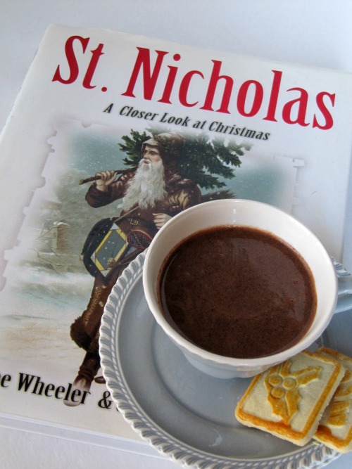 overhead view of a white cup of decadent dark hot chocolate on a white plate with two vanilla chookies sitting on a Christmas book about st. nicholas.