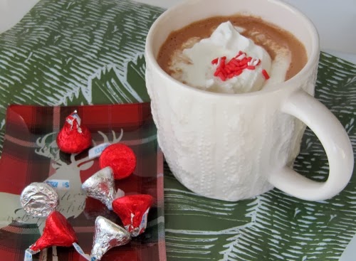 Side view of a large white mug of dark and milk chocolate homemade hot chocolate drink with whipped cream topping and a few red sprinkles sitting on a gray and white table cloth next to a red and white dish of Hershey's candy kisses.