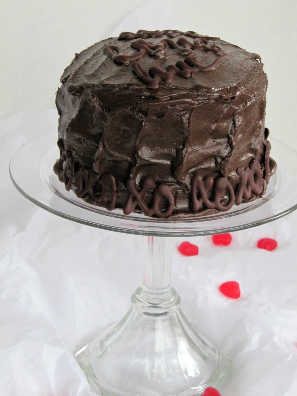A round fudge-filled chocolate cake frosted with dark glossy chocoate frosting sits on a footed glass cake stand with red Valentine decorations surrounding on white parchment paper