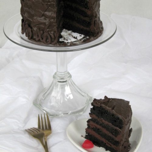 A fudge-filled dark chocolate layer cake frosted with Hershey's chocolate frosting sits on a glass cake stand with one piece of the cake on a heart shaped plate in front of the stand