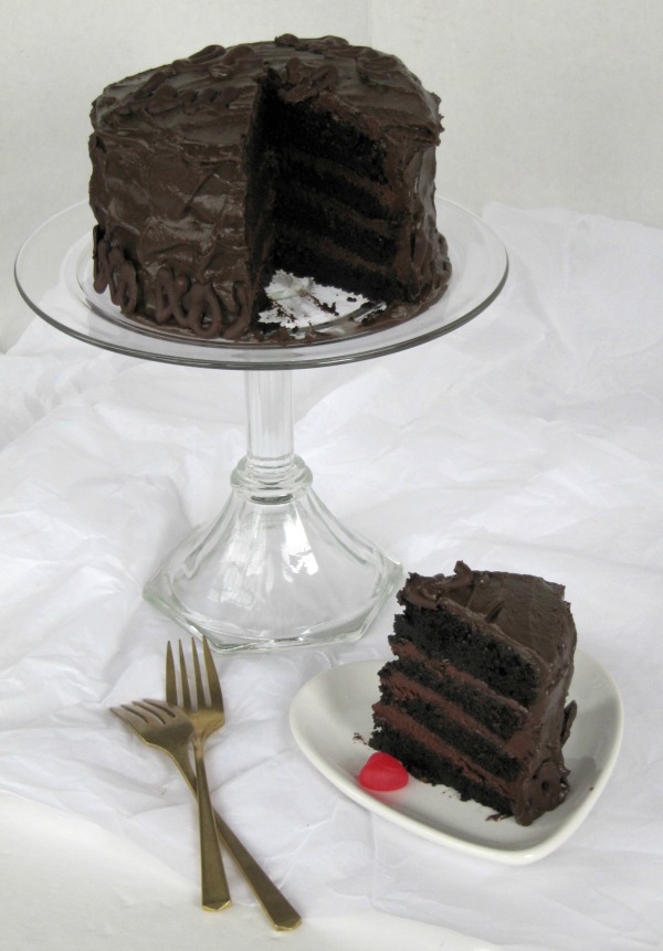 A chocolate cake with fudge filling and Hershey's chocolate frosting 
 for Valentines Day sits on a glass cake stand with one piece of the cake on a heart shaped plate in front of the stand.