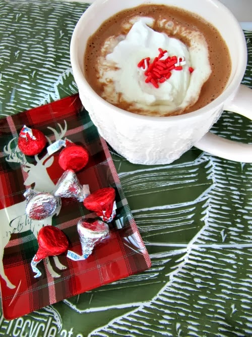 Top view of a steaming cup of homemade hot chocolate topped with whipping cream and a few red sprinkles, sitting on a gray and white cloth next to a tray of Hershey's candies