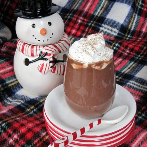 A stack of red-rimmed white dessert plates holds a tall glass of hot chocolate topped with eggnog-flavored whipped cream along with a white spoon with a candy-striped handle. A white ceramic snowman sits behind the plates on a red plaid table cloth