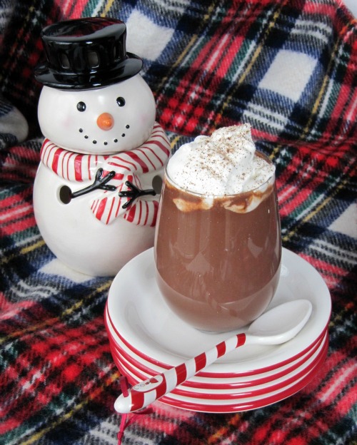 A stack of red-rimmed white dessert plates holds a tall glass of hot chocolate topped with eggnog whipped cream for hot chocolate along with a white spoon with a candy-striped handle. A white ceramic snowman sits behind the plates on a red plaid table cloth.