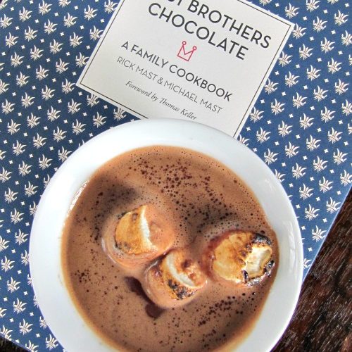 overhead view of a white cup of mast brothers classic hot cocoa topped with toasted marshmallows sitting on a blue and white mast brothers chocolate family cookbook