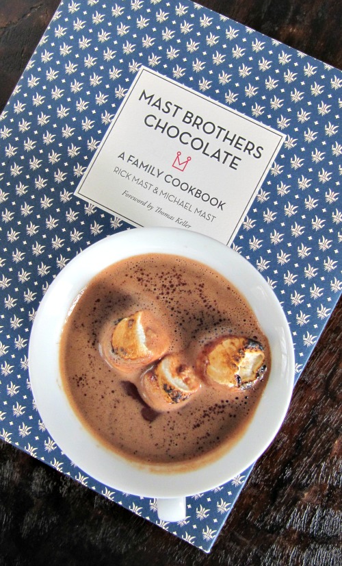 overhead view of a white cup of mast brothers classic hot cocoa topped with toasted marshmallows sitting on a blue and white mast brothers chocolate family cookbook
