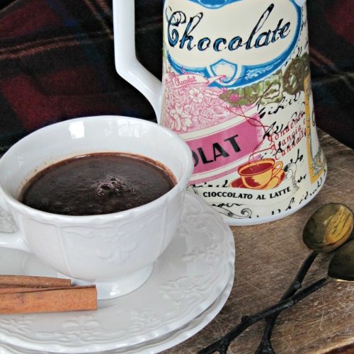 close up image of a white cup of rich hot mississippi mud pie cocoa made with chocolate, bourbon and spices. The cup is on a stack of two white saucers with to sticks of cinnamon in front of a colorful pitcher on a wood cutting board