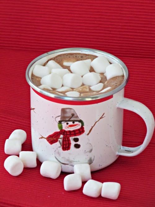Closeup view of mocha hot chocolate with marshmallows in a white cup with a snowman on it and several mini-marshmallows scattered on a red surface.