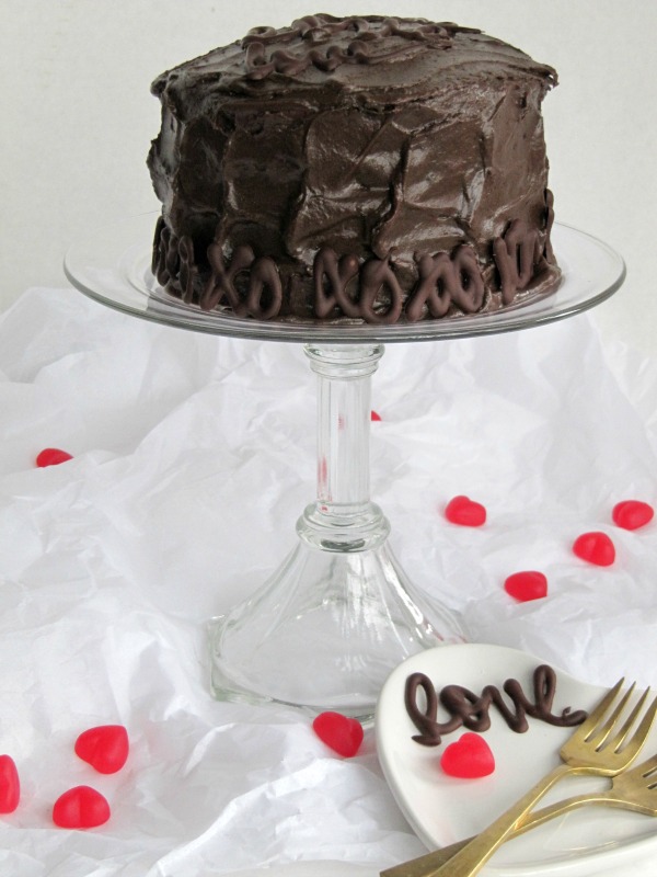 A large round chocolate cake with fudge fillin gand Hershey's chocolate frosting for Valentines Day sits on a tall glass cake stand with Valentine's decorations surrounding it against white parchment paper.