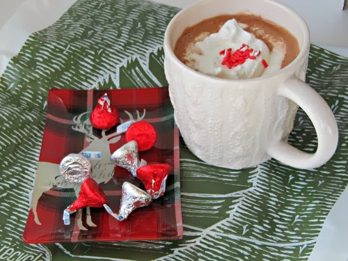 side view of a white ceramic handled mug of rich homemade hot chocolate made with three kinds of chocolate and cream, on a gray and white table covering next to a rectangular tray of christmas candies.