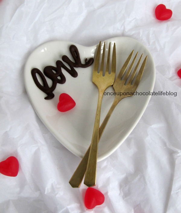 A white ceramic Valentine heart-shaped dish with chocolate candy love decoration, red candy hearts and two forks sits on white parchment.
