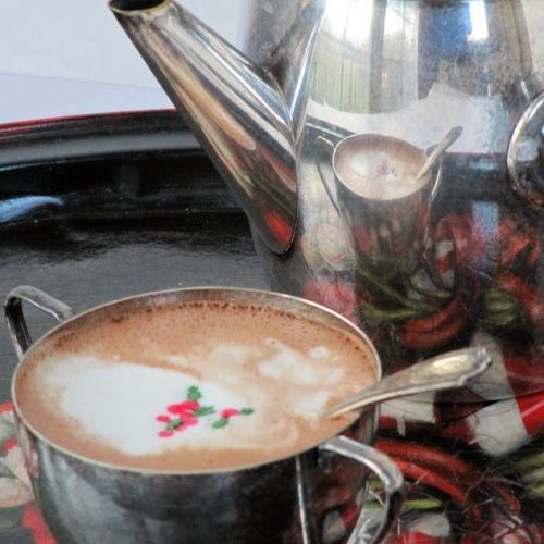 Silver cup of wittamer hot chocolate with christmas sprinkles on top is sitting on a christmas platter on a dark tray next to a tall silver pot