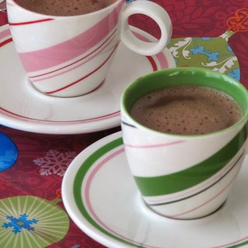 Two peppermint striped cups and saucers, one pink and white, one green and white, both filled with Bicerin coffee and chocolate drink. The cups are on a colorful table cloth.