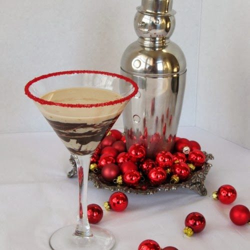 A side view of a tall martini glass with red sprinkles around the edge that holds a Bailey's chocolatini cocktail made with vodka, Bailey's and chocolate liqueur standing in front of a silver snowman martini shaker surrounded with small red christmas ball decorations.