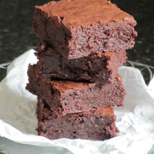 A cloeup of a stack of four fudgy Baked Bakery NY aka Oprah's favorite brownies are sitting on a piece of white parchment against a dark background.