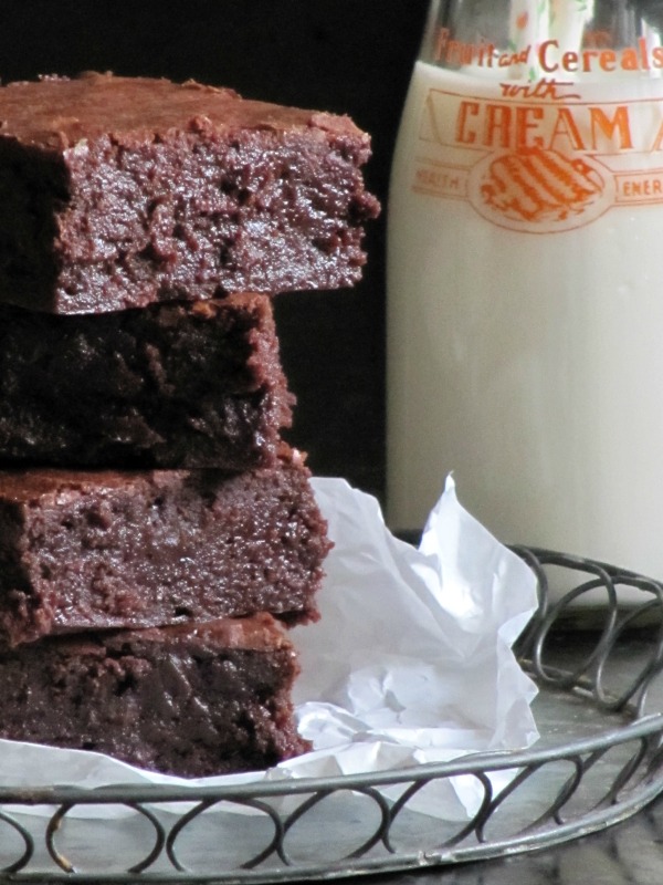 A closeup view of four moist dark chocolate Baked Bakery NY aka Oprah's favorite brownies on white parchment on a round wire tray in front of a glass milk bottle full of milk.