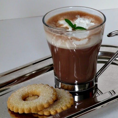 a silver tray on a gray surface holds a tall glass mug of basil mint hot chocolate with whipped cream and a sprig of fresh mint along with two round sugar cookies