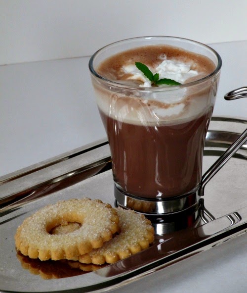 a silver tray on a gray surface holds a tall glass mug of basil mint hot chocolate with whipped cream and a sprig of fresh mint along with two round sugar cookies