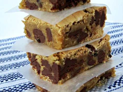 A closeup image of a stack of four triple chocolate blondie brownie bars separated with parchment squares with each bar showing a rich chewy center abundant  with chocolate chip. The stack is on a blue and white striped table cloth. 
