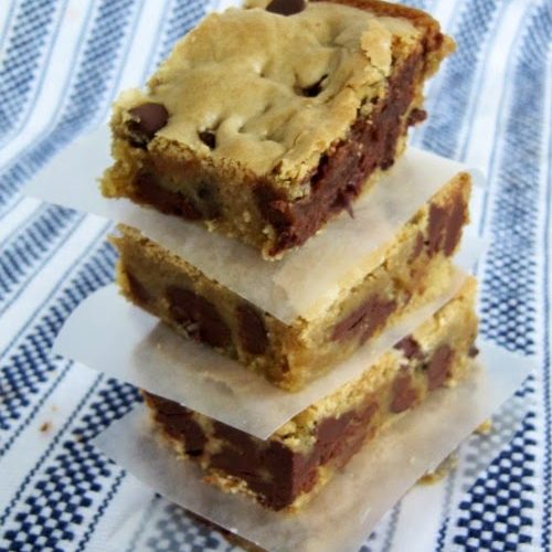 An angled overhead view of a stack of triple chocolate blondie bar cookies with crispy tops and chocolate chip-filled centers on a navy and white striped woven table cloth.s
