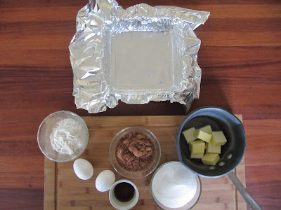 Ingredients for making Browned Butter Brownie Recipe from Bon Appetit, like butter, eggs, sugar, flour and chocolate, plus a foil-lined baking pan to cook the brownies all sitting on a cutting board on a wood table.