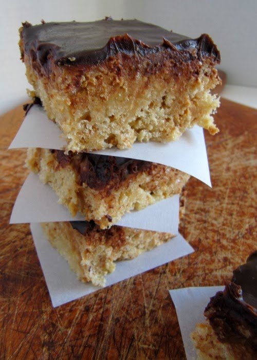 A stack of browned butter peanut butter cup rice krispies treats with chocolate ganache topping is sitting on a brown surface with white parchment separating the squares and a single square on a piece of parchment is in front.