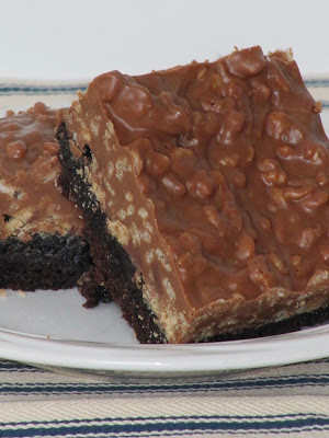 A white plate holding two peanut butter cup crunch brownies with a brownie base and a thick peanut flavored layer on top with rice krispie cereal and peanuts.