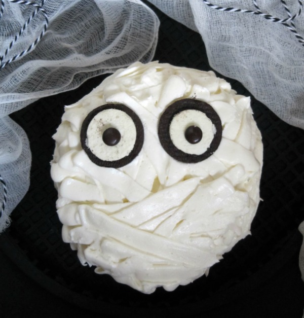 A closeup of a chocolate mummy cake for your halloween celebrations made with chocolate cake frosted with vanilla buttercream to look like bandages with two halved Oreos for eyes and two plain brown M&M candies for eye pupils. The cake is on a dark table surrounded with white cheesecloth and blue and white string decorations.