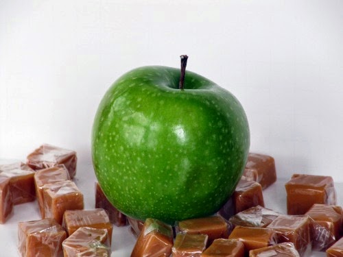 A shiny green apple is surrounded with Kraft caramels wrapped in plastic sitting on a white surface against a g=white background ready for making chocolate caramel apples.