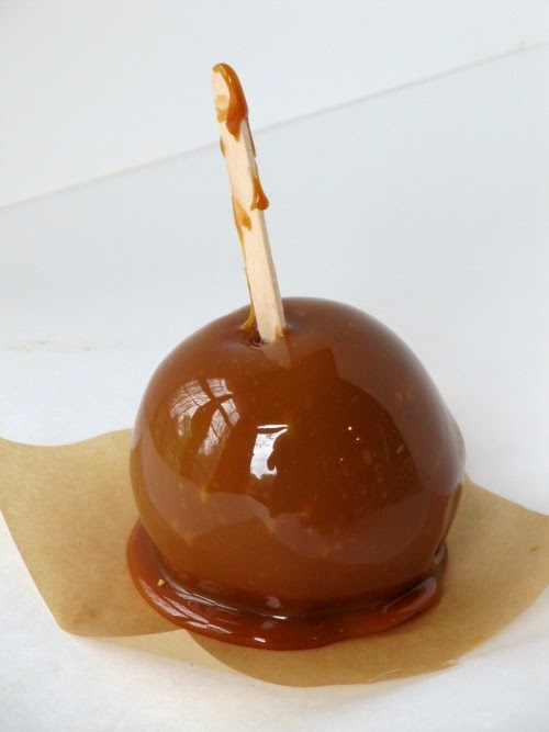 A single caramel covered apple with a wooden popsicle stick is sitting on a piece of light brown parchment paper, ready to become a chocolate caramel apple.