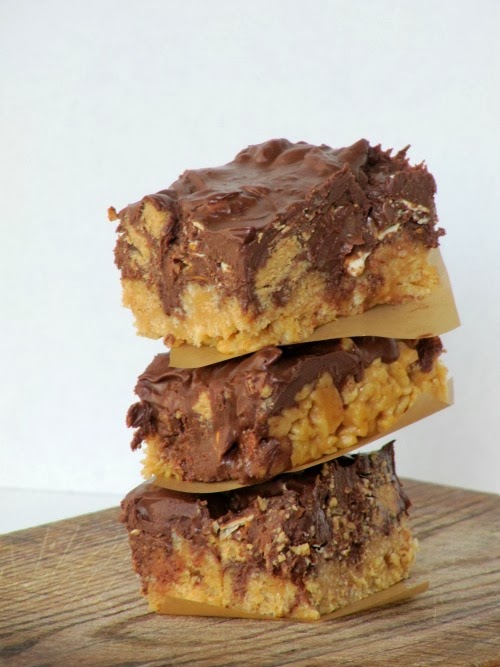 A stack of three luscious peanut butter pretzel bars with melted chocolate chip and pretzel topping and made with rice krispie cereal is sitting on a wood surface.