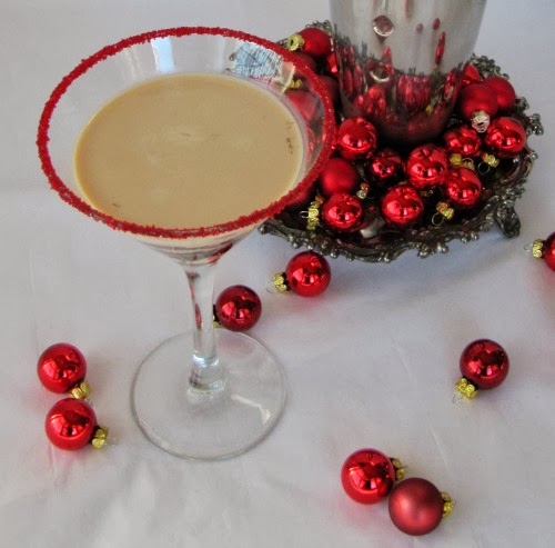 A closeup image of a Bailey's chocolatini in a martini glass with red sprinkles aruond the edge in front of little red Christmas ball decorations.