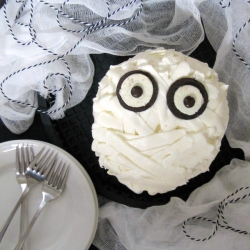A chocolate mummy cake for your Halloween celebrations, frosted with white buttercream to look like bandages and two open Oreos and two brown M&M candies for eyes, is sitting on a dark table surrounded with a stack of white dessert plates and forks and some white cheesecloth and blue and white string decorations,