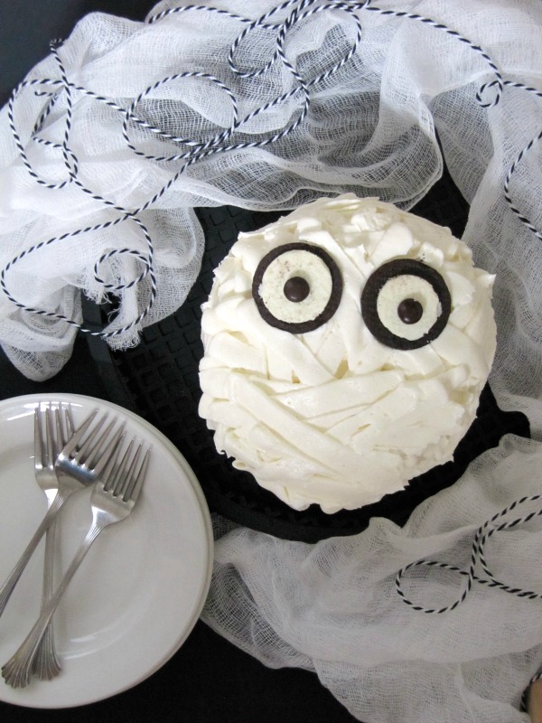 A chocolate mummy cake for your Halloween celebrations, frosted with white buttercream to look like bandages and two open Oreos and two brown M&M candies for eyes, is sitting on a dark table surrounded with a stack of white dessert plates and forks and some white cheesecloth and blue and white string decorations,