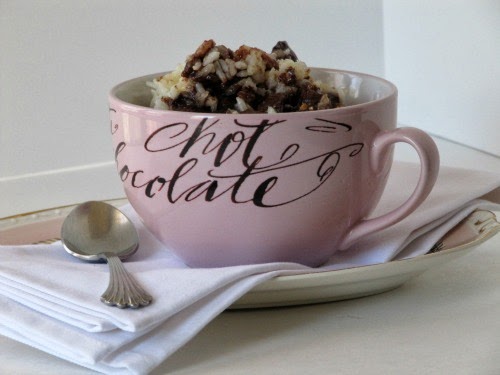 A side view of a large pink round mug holding a serving of baked chocolate almond toffee rice pudding. The cup is on a white napkin on a plate along with a silver spoon.