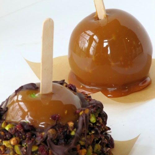A caramel apple and a chocolate caramel apple are both sitting on pieces of brown parchment paper on a white surface, with the chocolate covered caramel apple having been rolled in chopped pistachios and dried cranberries.