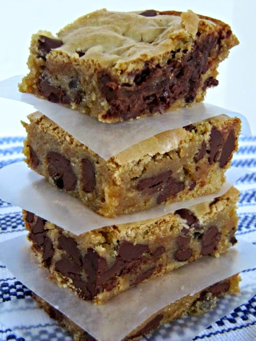 A side view of a tall stack of rich triple chocolate blondies chock full of dark, milk, and semi-sweet chocolate chips with crunchy tops. The stack is sitting on a blue and white table covering against a white background.