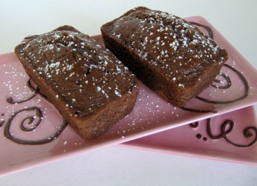 A side view of two light pink rectangular trays balanced on each other and holding two whole loaves of quadruple chocolate bread dusted with shaved white chocolate.