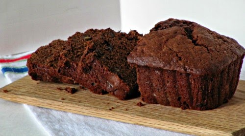 Side view of a half sliced loaf of chocolate chocolate chip banana bread on a light brown cutting board on a white table cloth.