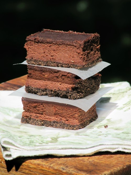 A side view of three stacked chocolate chocolate cheesecake bars with white parchment in between them are sitting on a light colored cloth napkin on a wood table against a black background.