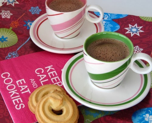 Against a colorful background including a cookie cookbook, two christmas peppermint cup and saucer sets, one pink and white and one gren and white, are filled with the Italian Bicerin chocolate and coffee drink along with two sugar cookies at the side.
