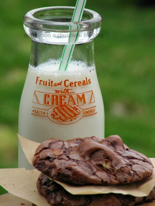 A stack of round chocolate reese's peanut butter egg cookies is sitting in front of a tall glass milk bottle filled with milk and a straw in front of a green background.